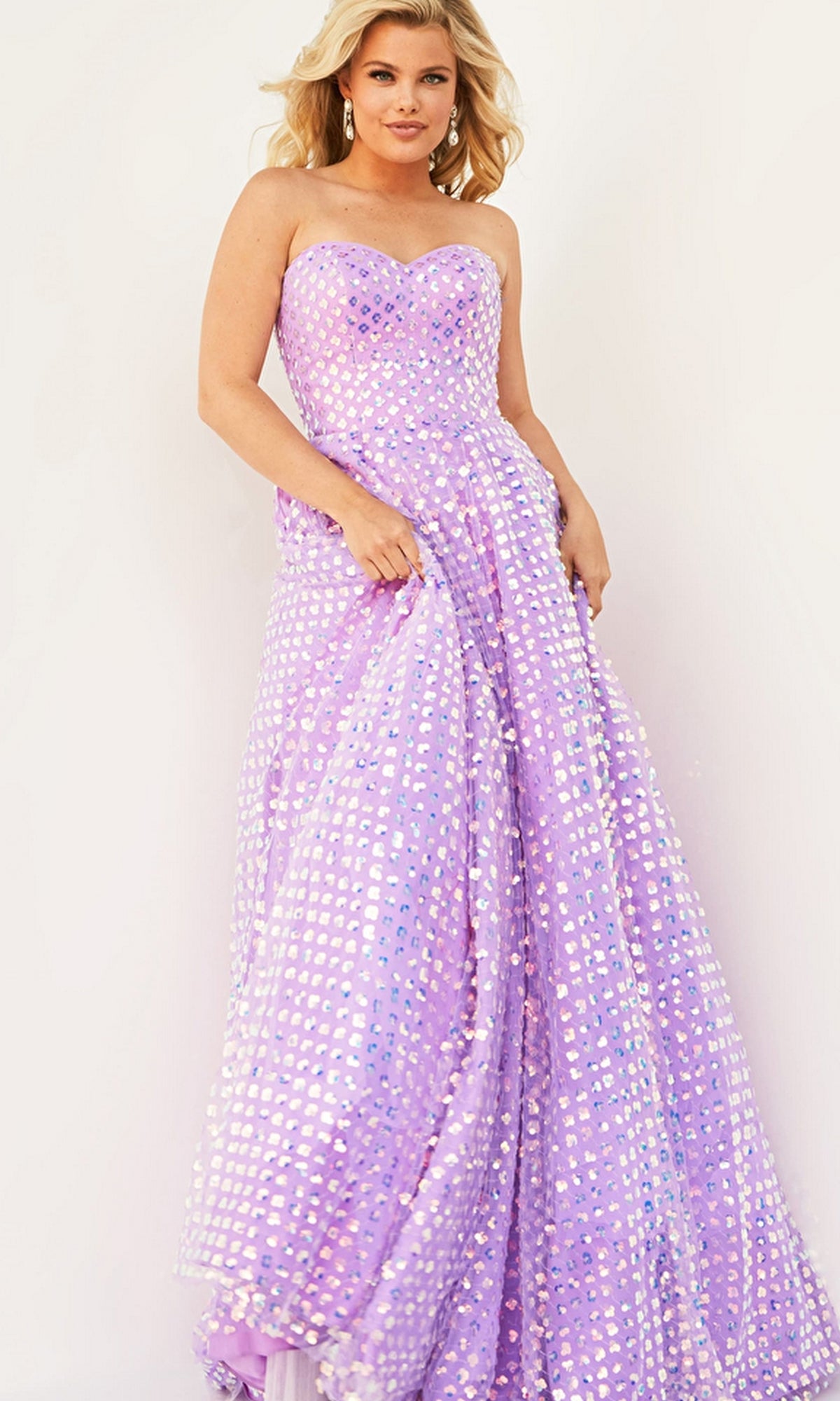 A woman wears the Jovani Plus-Size Strapless Long Prom Dress 08605B, a lilac gown featuring a fitted bodice and full skirt adorned with shiny sequin-like embellishments, as she stands against a plain light background.